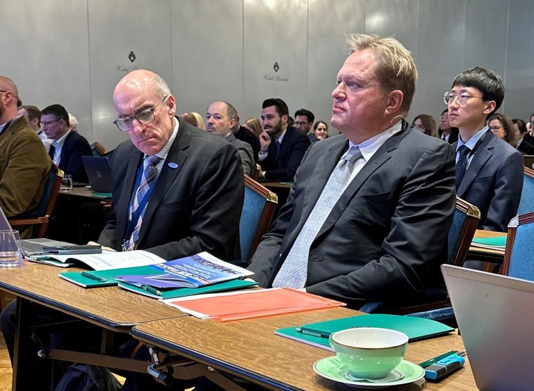 Two men in suits listening to a lecture. More people in the background.