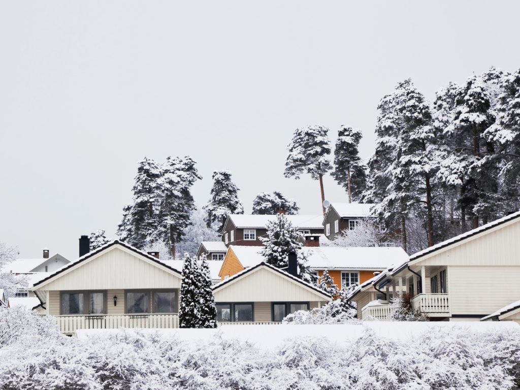 Vinterlandskap med flere eneboliger bak hverandre. Snø på bakken og på husene. Store furutrær i bakgrunnen med snø på.